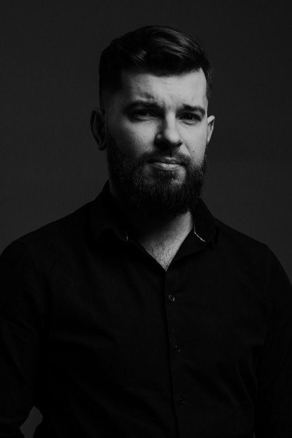 Czarno-biały portret mężczyzny z brodą w ciemnej koszuli, na ciemnym tle.