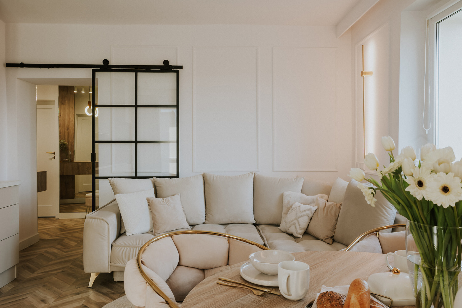 Jasna sofa i stół z kwiatami przy przesuwnych drzwiach z czarnymi szprosami.